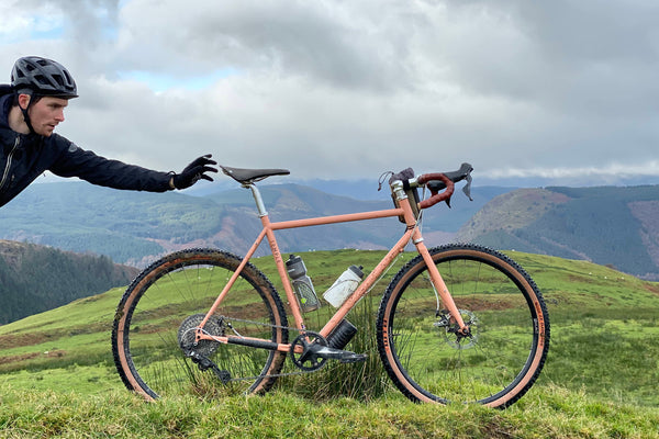 Temple Cycles pale orange bike on a mountain scape with a man in a helmet reaching in from the left hand side
