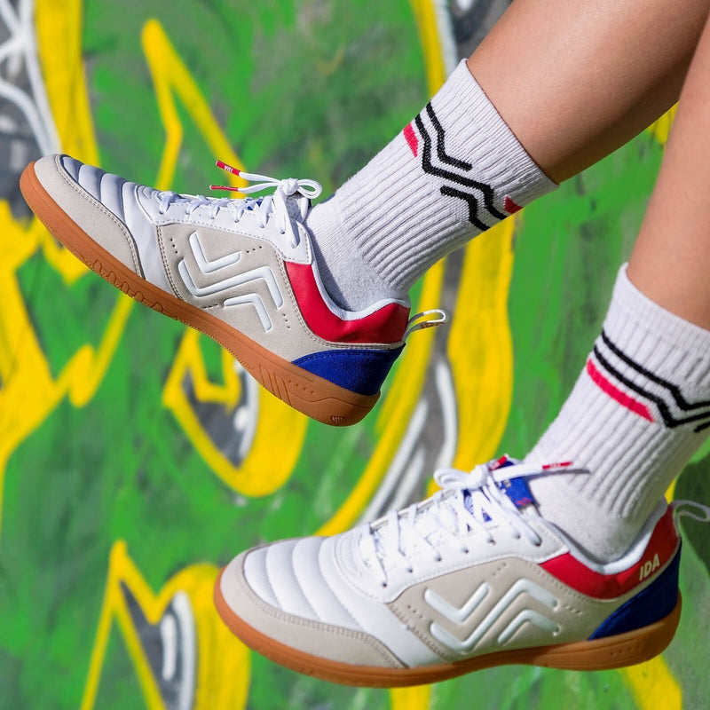 IDA Spirit Women's Indoor Soccer Futsal Shoe, White with Red & Blue accents, IC, Indoor Court, photo of person wearing them with white socks and dangling their feet in front of a green and yellow graffiti background 