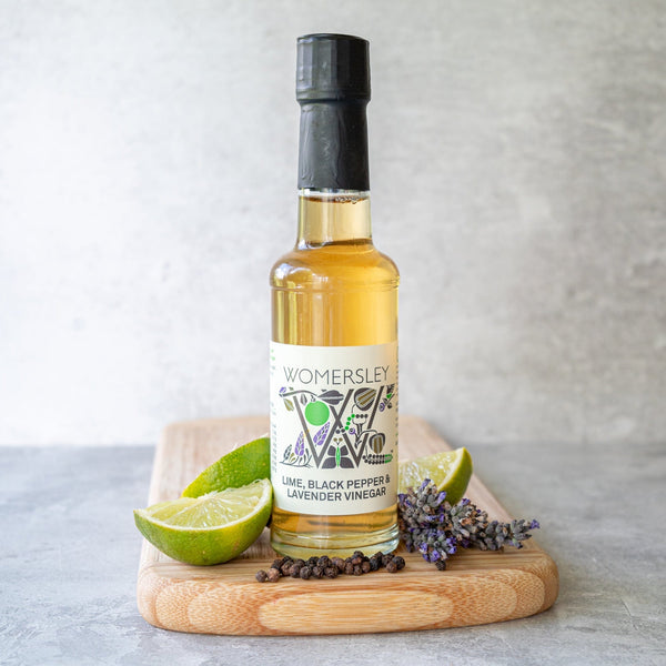 Womersley Foods 150ml Lime, Black Pepper & Lavender Fruit Vinegar standing on a cutting board with grey background surrounded by Lime, Black Pepper & Lavender ingredients on a grey background.