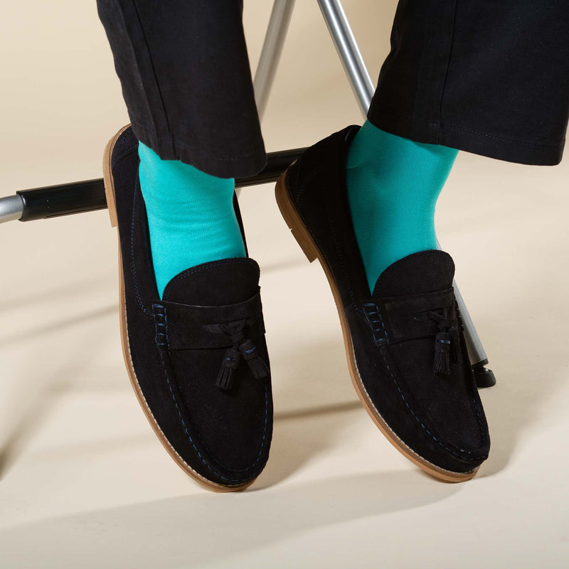 Man sitting on stool wearing black boat shoes, black trousers and turquoise Classic men's luxury socks